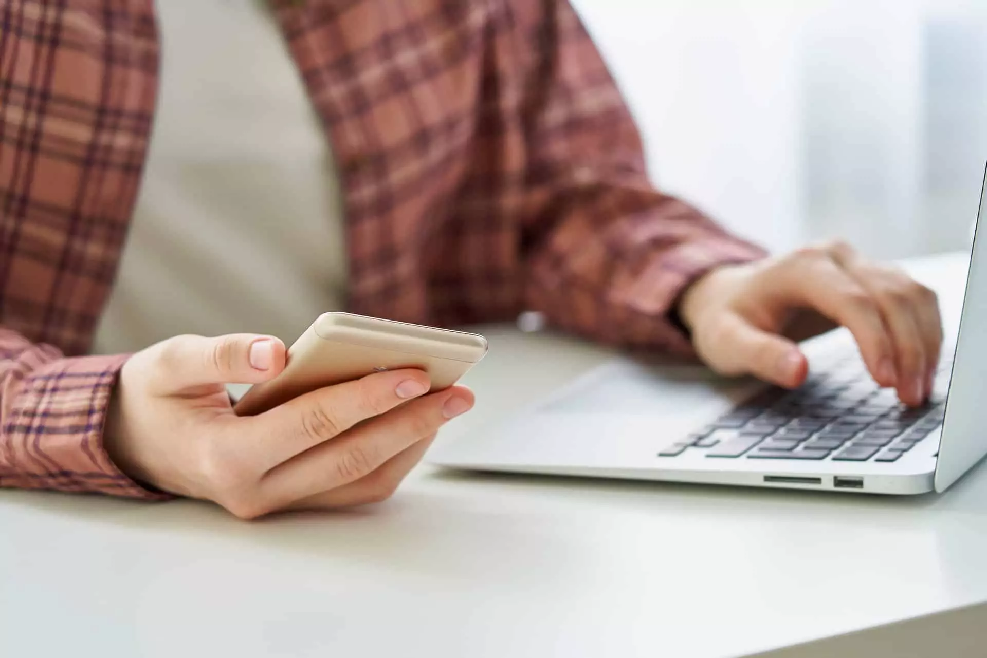 Faceless young female browsing smartphone while typing on laptop. Millennial data privacy concept